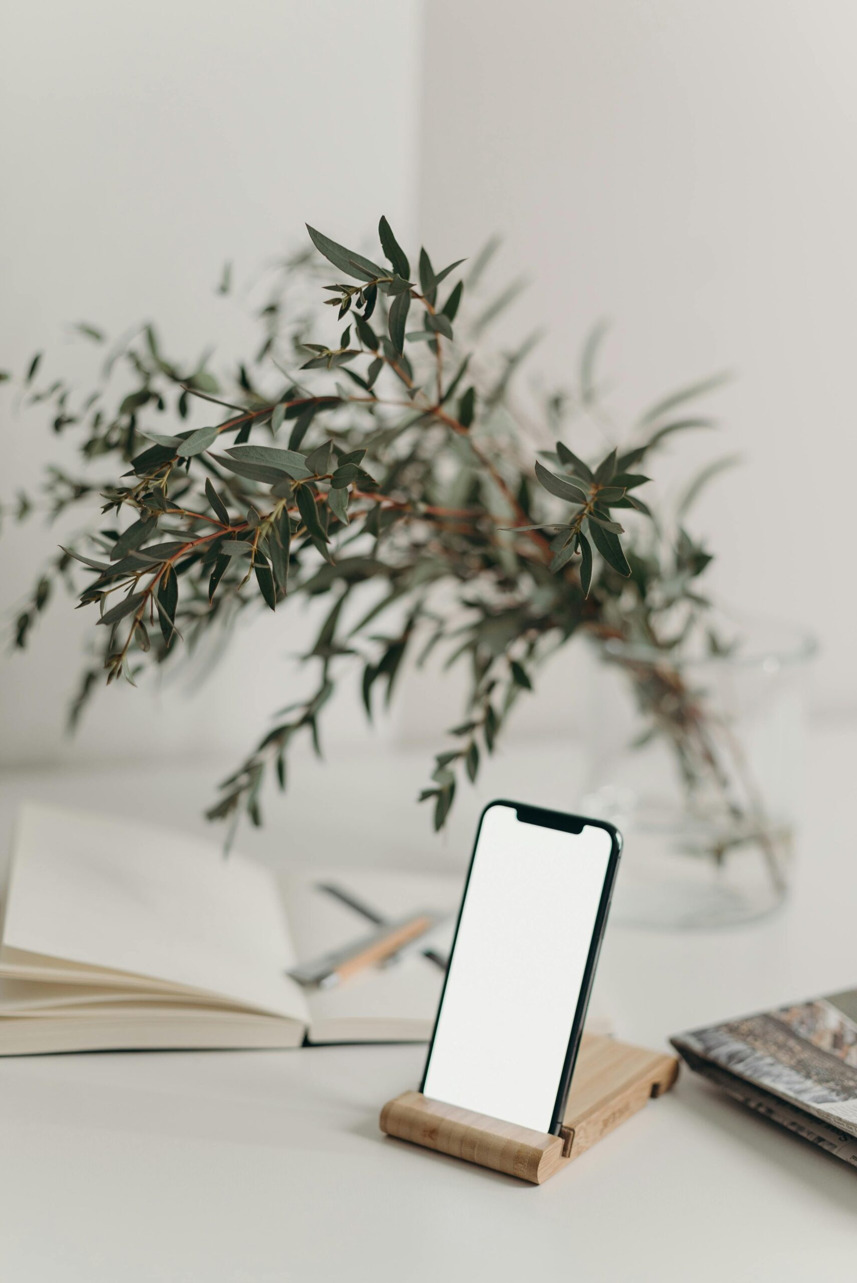 A sleek smartphone with a blank screen on a wooden stand, styled with a decorative plant and open notebook.